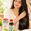 A woman with long, shiny black hair is shown indoors beside a bottle and box of Al Haque's Amla Pure Hair Oil on a wooden table, with green amla fruits and a towel nearby.