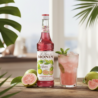 Monin Guava syrup bottle with a glass of guava juice on a wooden table with guava slices and leaves.
