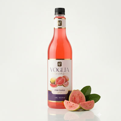 A bottle of Voglia Pink Guava syrup with a black cap, shown next to fresh pink guava slices and green leaves on a clean white background.