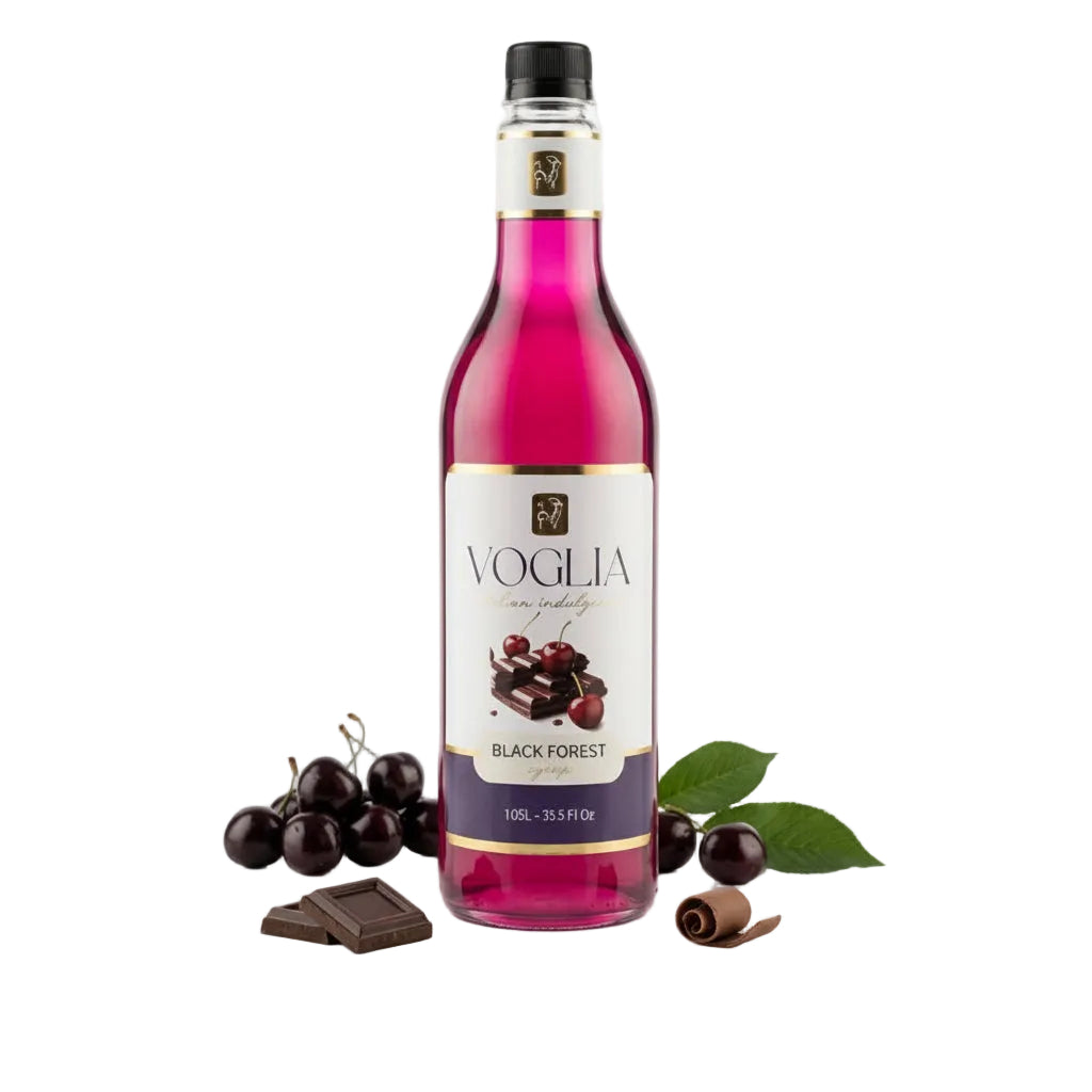 A bottle of Voglia Black Forest syrup with a vibrant magenta-pink liquid, styled with fresh cherries, chocolate pieces, and leaves on a reflective white surface.