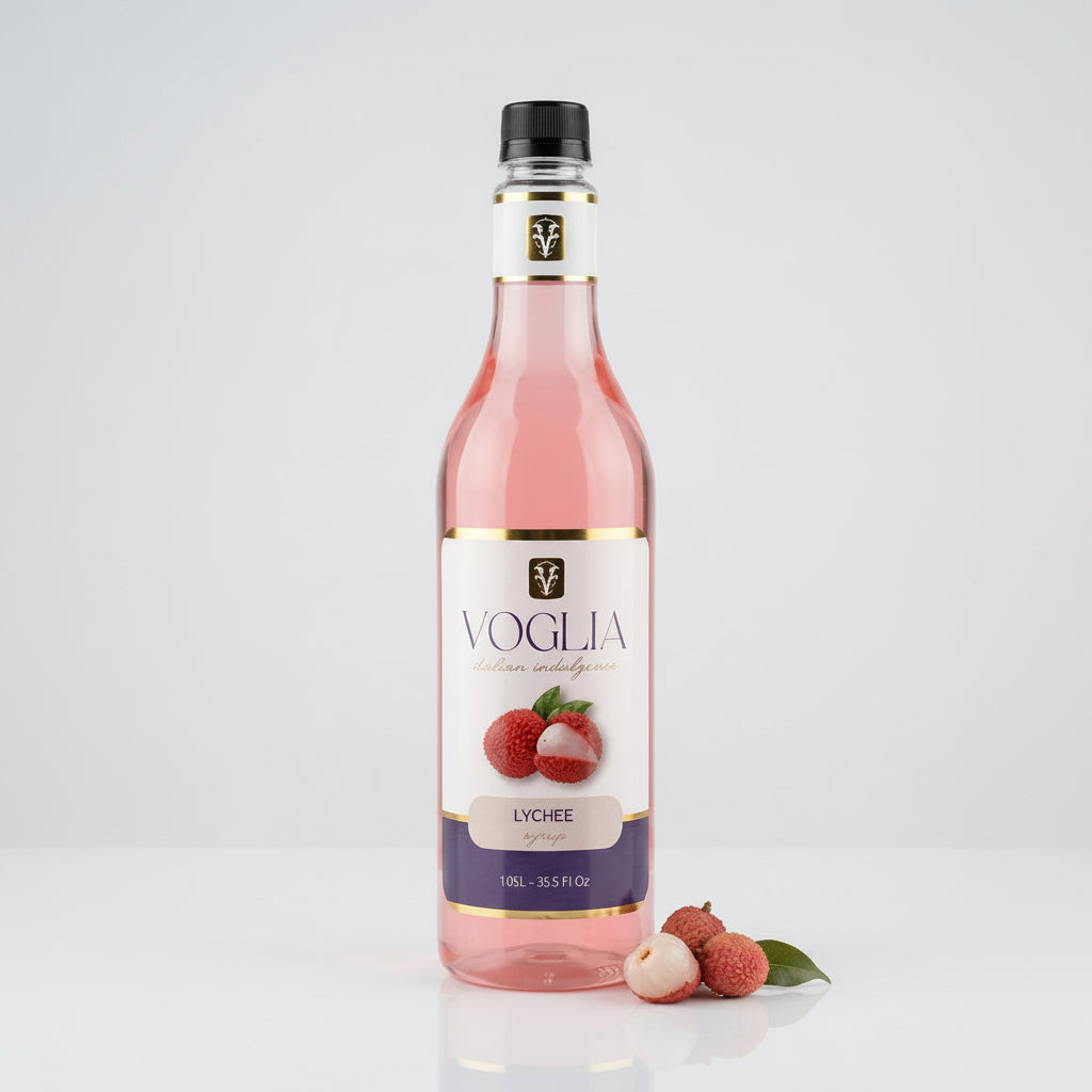 A bottle of Voglia Lychee syrup filled with pink liquid, standing on a white reflective surface with lychee fruits placed beside the base. The bottle has a black cap, a white label with gold trim, and a purple band near the bottom.