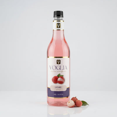 A bottle of Voglia Lychee syrup filled with pink liquid, standing on a white reflective surface with lychee fruits placed beside the base. The bottle has a black cap, a white label with gold trim, and a purple band near the bottom.