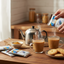 Person pouring a packet of instant coffee into a cup on a wooden table with cookies and a teapot.