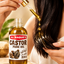 A bottle of Al Haque's Castor Pure Oil sits on a stone surface while a person holds a glass dropper above their dark hair, showing golden oil dripping onto the strands. The background is a soft, warm beige indoor setting.