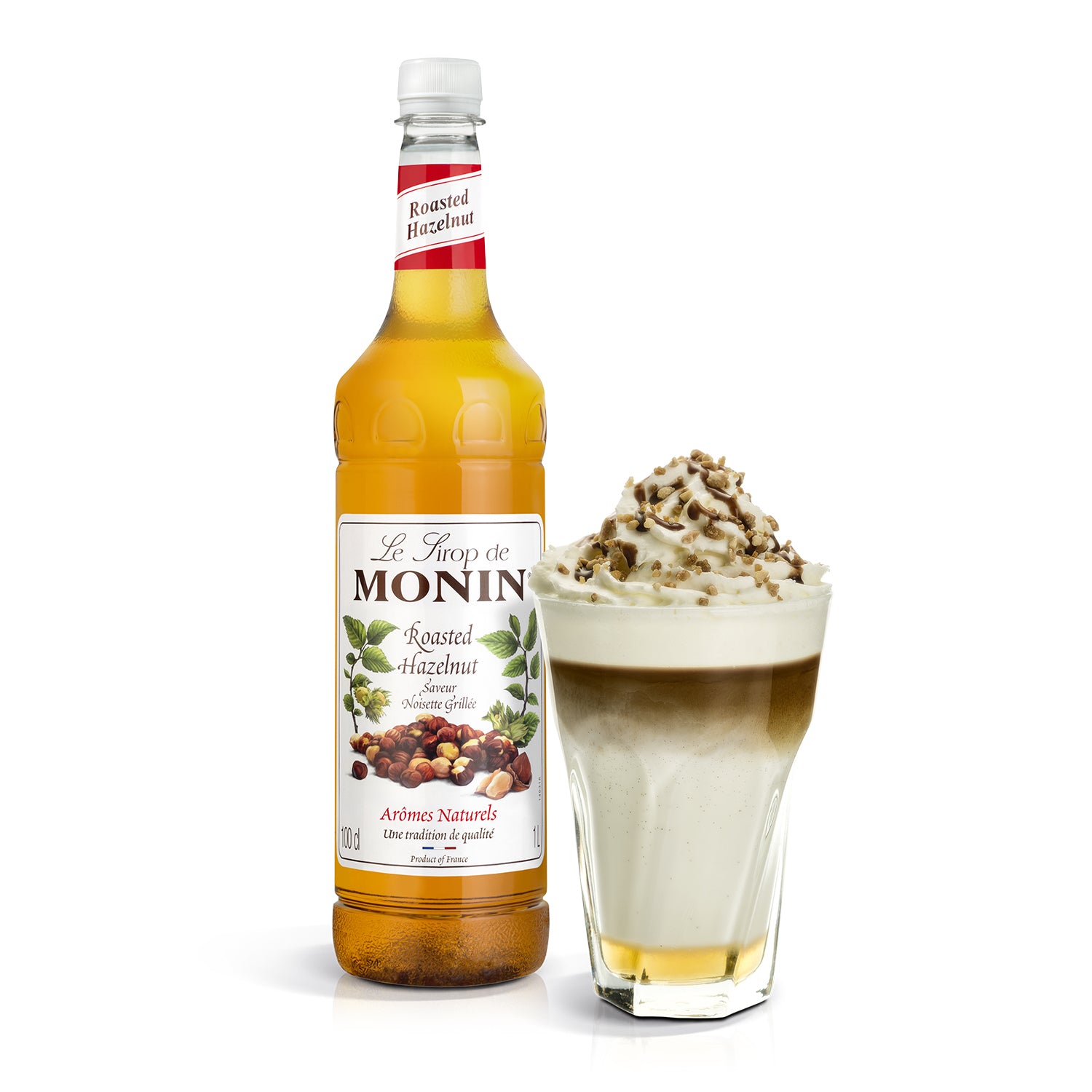 Monin hazelnut syrup bottle next to a glass of a layered dessert with whipped cream and nuts.