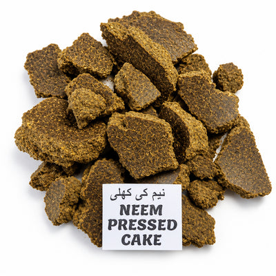 A pile of dark brown, textured pressed cake pieces with a small white label reading 'NEEM PRESSED CAKE' in English and Urdu, set against a solid white background.